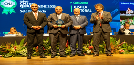 A foto mostra quatro homens de terno em pé no palco de um auditório, sorrindo. O homem ao centro...