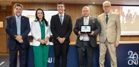 Cinco pessoas estão em pé lado a lado em um auditório, posando para foto oficial. Uma delas segu...
