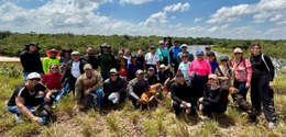 Grupo grande de pessoas posa ao ar livre em área de vegetação baixa, às margens de um rio, sob c...