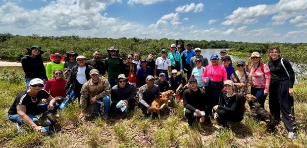 TRE-RR- Trilhas Ecológicas Grupo grande de pessoas posa ao ar livre em área de vegetação baixa, às margens de um rio, sob c...
