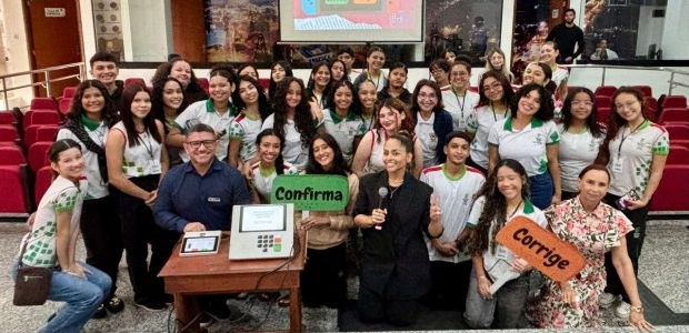 Grupo de estudantes posa sorrindo em um auditório com servidores do TRE-RR. À frente, há uma urn...