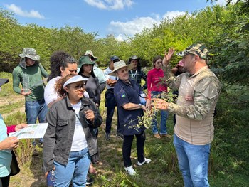 Grupo de pessoas caminha por uma trilha cercada de vegetação, em área sombreada por árvores. Tod...